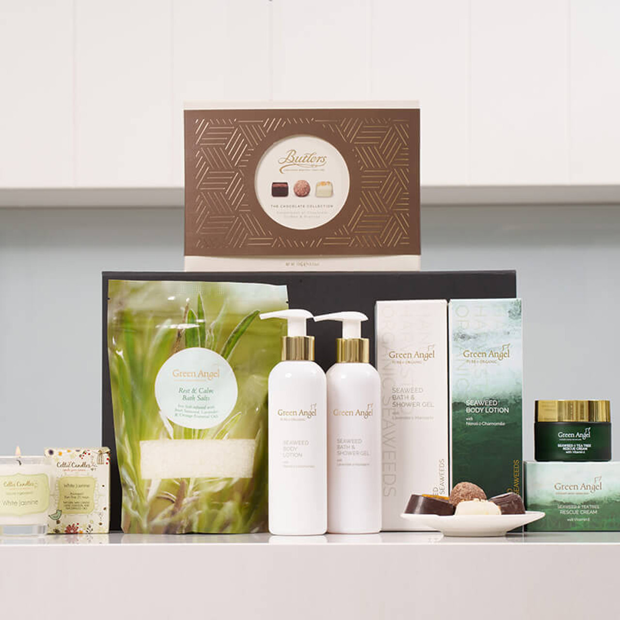 Ultimate Relaxation Pamper Gift: A collection of skincare and personal care products is displayed on a shelf. The set includes two white bottles of lotion, various boxes and jars with green packaging, and a small candle. A gift box, prominently featured at the back, contains assorted treats.