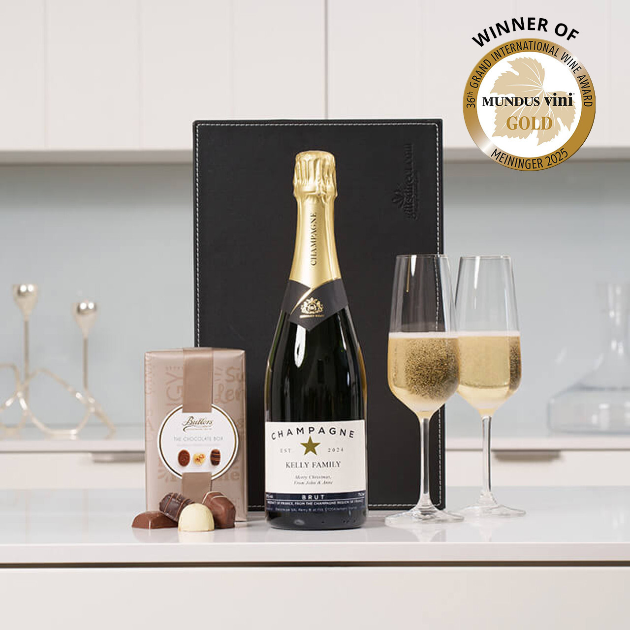 A bottle of champagne with a personalised label is placed on a countertop beside two fluted glasses filled with sparkling champagne. Next to the champagne is a box of  of Butlers chocolates, with some chocolates displayed outside the box. A black gift box is partially visible in the background. In the corner, there is a seal indicating a gold award from Mundus Vini.