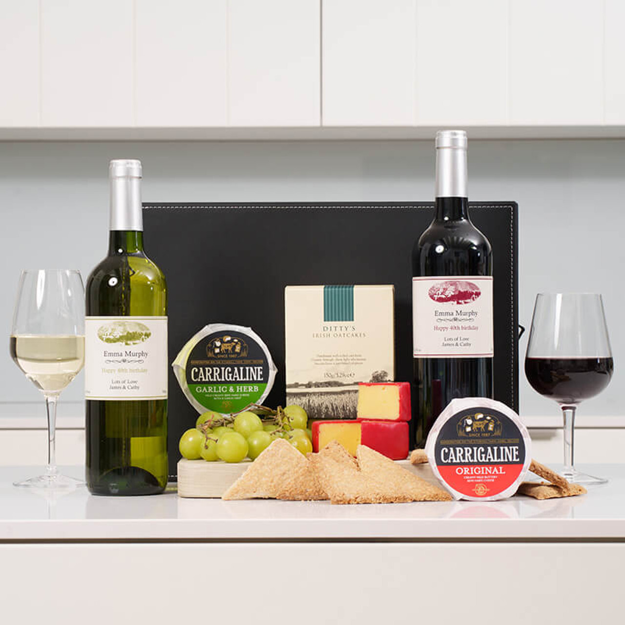 Two bottles of wine, a white and a red with personalised labels , are placed on a table alongside two glasses filled with their respective wines. In the foreground, there is an assortment of cheeses, crackers, and grapes. The background features a simple countertop.