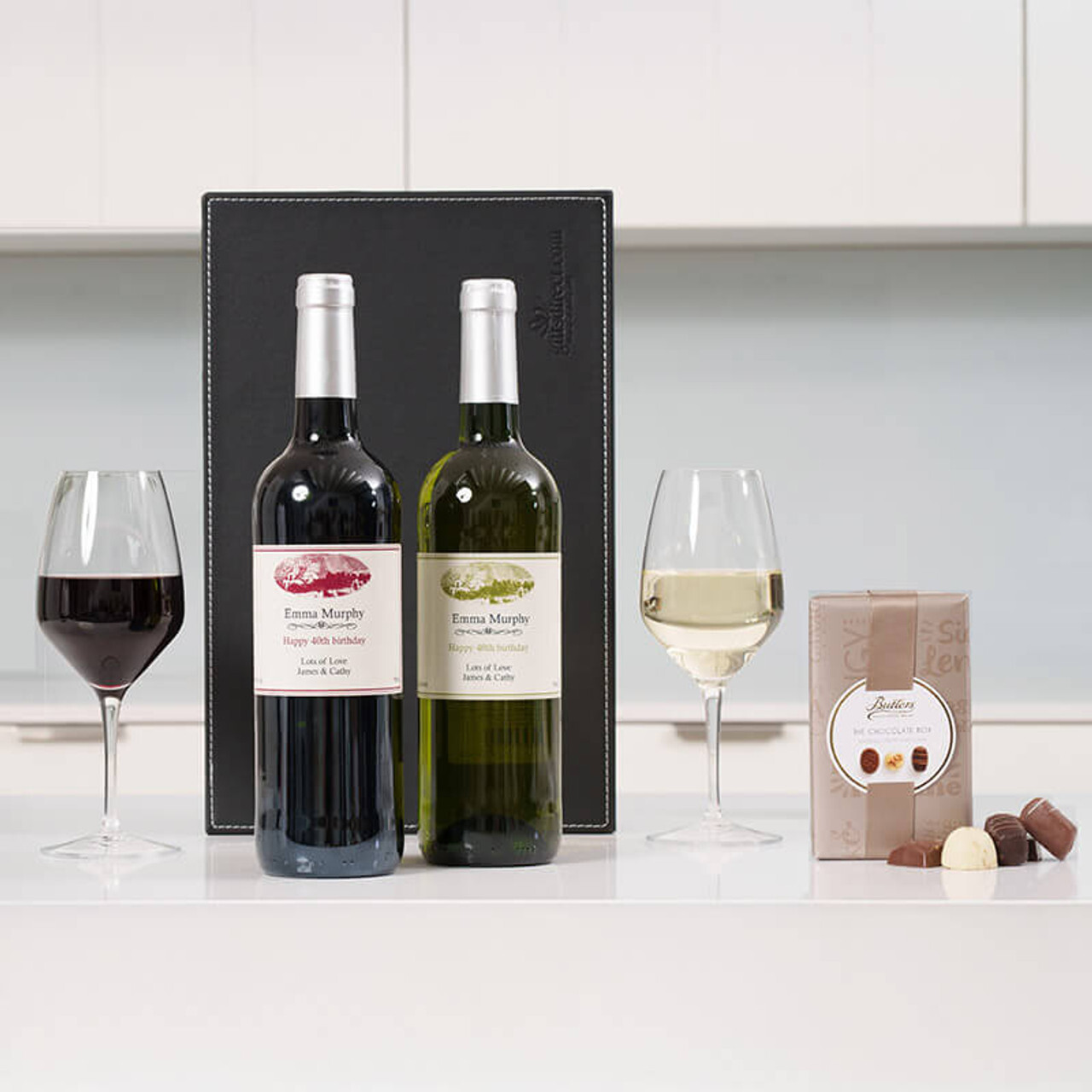 Personalised Wine Duo & Chocolates. Two bottles of wine, one red and one white with personalied labels, are placed next to two wine glasses filled with their respective types. A box of chocolates sits nearby, all set against a minimalist kitchen background.