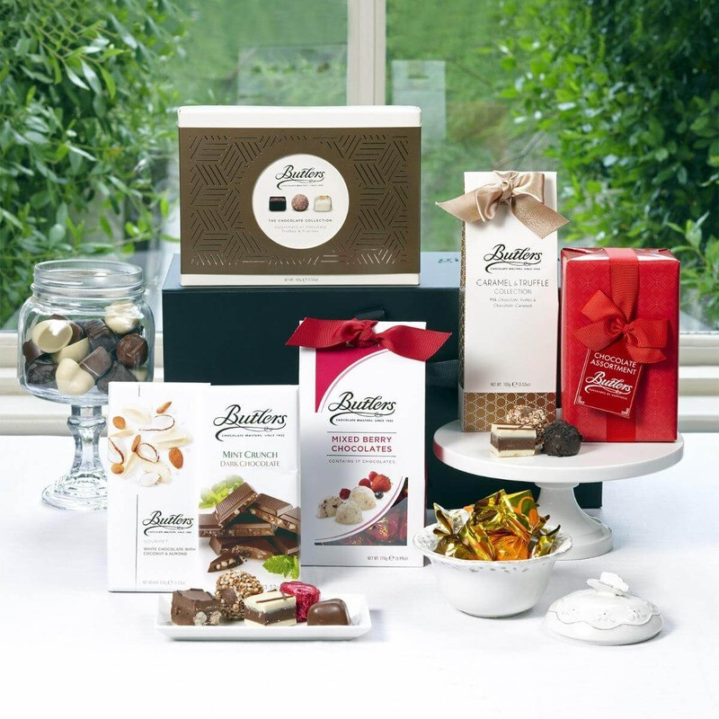 Valentine's Luxury Butlers Chocolate Gift Box: An assortment of chocolates and confectionery items is displayed on a table. Various packages from Butlers Chocolate include boxes with different flavours, a glass jar filled with assorted chocolates, and decorative ribbons. The setting features greenery in the background, suggesting a bright and inviting atmosphere.