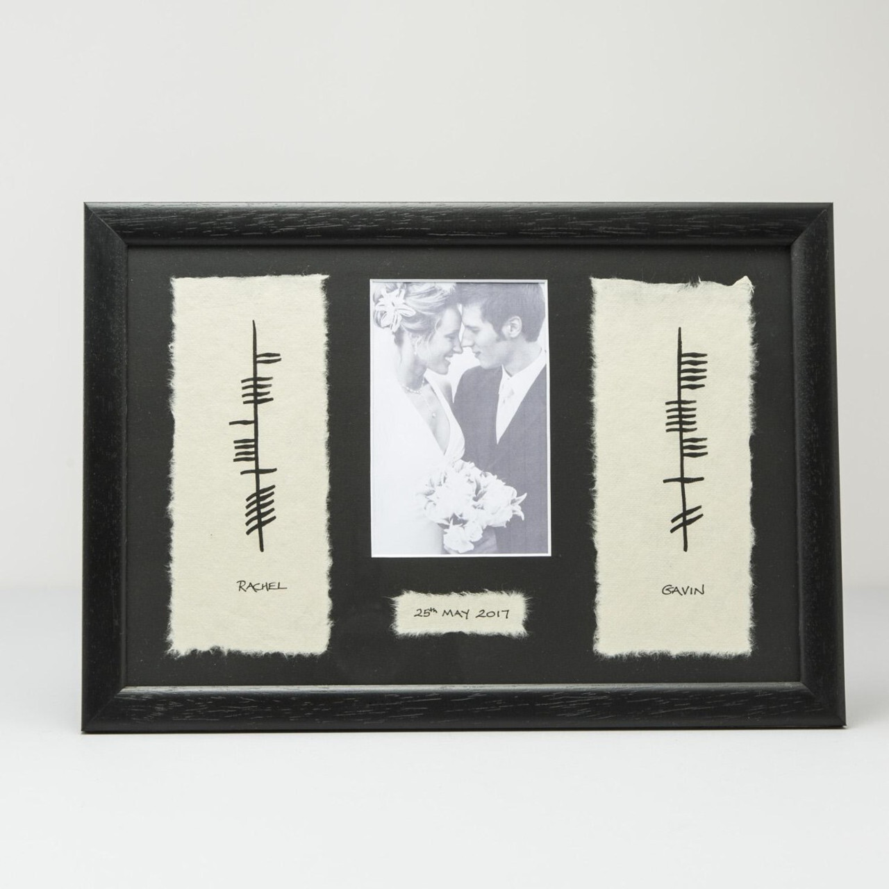 A black-framed photo display featuring a central black-and-white image of a couple on their wedding day, surrounded by two textured white panels. Each panel has Ogham writing and includes the names 