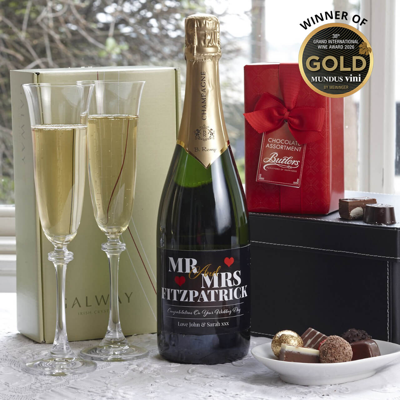 Personalised Mr & Mrs Wedding Gift Box & Champagne Flutes. A bottle of champagne labeled "MR & MRS FITZPATRICK" is displayed prominently beside two filled champagne flutes and a red box of chocolates from Butlers. A gold award seal indicating a win at the Mundus Vini competition is visible in the top corner. The scene is set near a window with soft natural light.