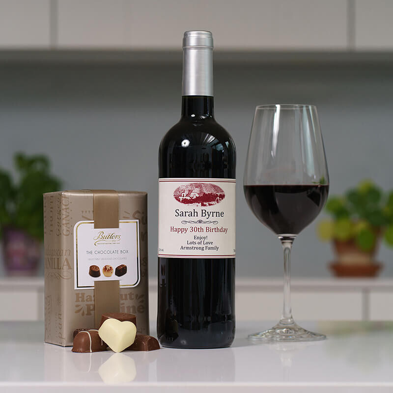 Personalised Red Wine & 160g Butlers Chocolates. A personalised bottle of red wine labeled "Sarah Byrne" next to a glass filled with the same wine. A box of chocolates is placed nearby, with one chocolate shaped like a heart in front. The background suggests an indoor setting with plants visible.