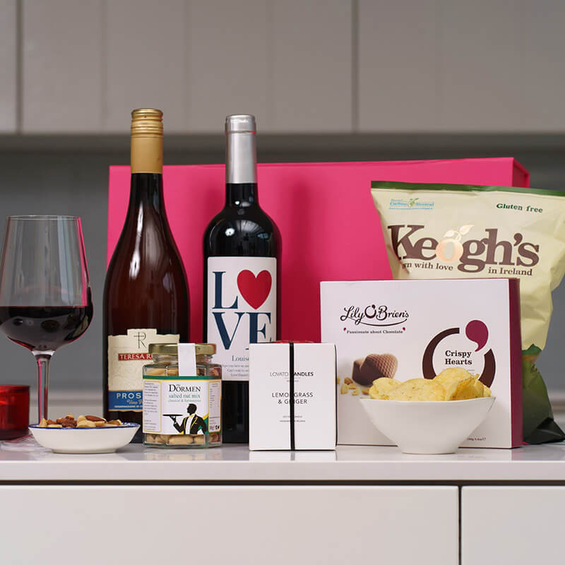 Romantic Night In Hamper. A countertop displays a selection of gift items. There are two bottles of wine, one with a label that reads "LOVE." A glass of red wine is filled beside the bottles. Additionally, there are gourmet snacks, including potato chips in a bag labeled "Keogh's," a box of chocolates from "Lily O'Briens," and a small jar with a decorative label. The background features a pink gift box and a minimalist kitchen setting.Romantic Night In Hamper. A countertop displays a selection of gift items. There are two bottles of wine, one with a label that reads "LOVE." A glass of red wine is filled beside the bottles. Additionally, there are gourmet snacks, including potato chips in a bag labeled "Keogh's," a box of chocolates from "Lily O'Briens," and a small jar with a decorative label. The background features a pink gift box and a minimalist kitchen setting.